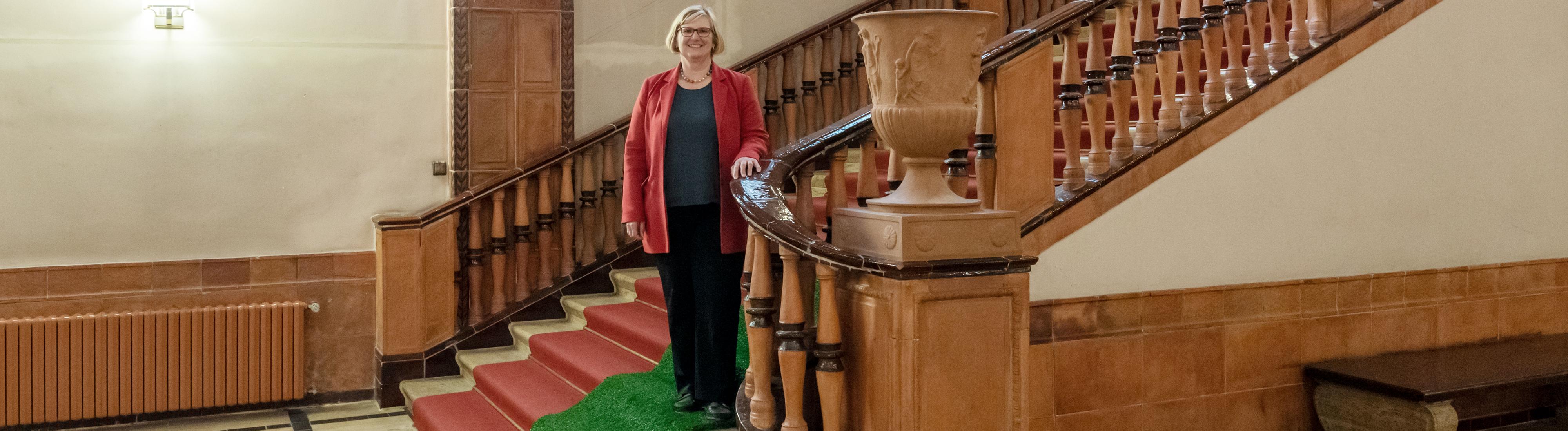 Portrait von Angelika Schöttler. Sie steht auf der marmornen Innentreppe des Schöneberger Rathauses, unter ihren Füßen ist ein grüner Teppich aus ökologischem Kunstrasen ausgerollt. Frau Schöttler trägt einen roten Blazer und eine Kette mit Kugeln in den Farben des Regenbogens.