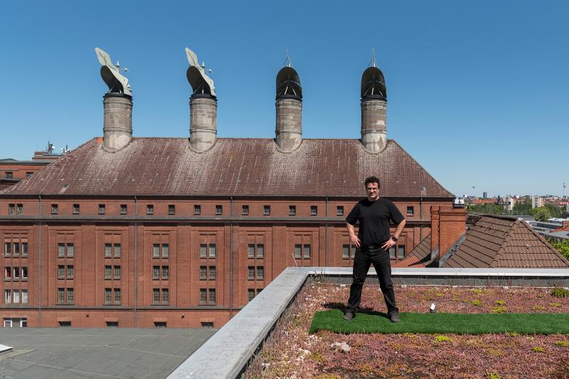 Portrait von Carsten Bredow auf dem Dach eines der Gebäude auf dem Gelände der Malzfabrik. Unter seinen Füßen ist ein Stück grüner Teppich und er hat ein Bein auf das Dachsims gestellt. Im Hintergrund ist das Hauptgebäude mit den vier Darren.