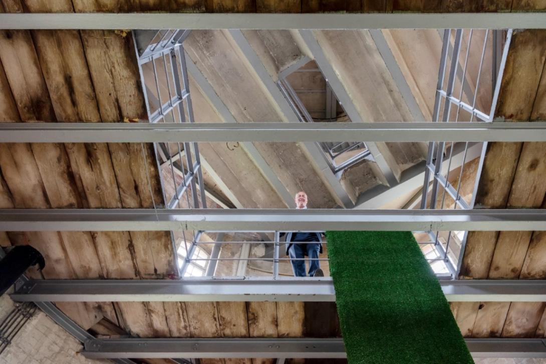 it von Guido Schütte im Businessanzug. Er ist im Obergeschoss des alten Wasserturms vom Boden aus fotografiert. Über dem Geländer neben ihm hängt der grüne Teppich aus nachhaltigem Kunstrasen.