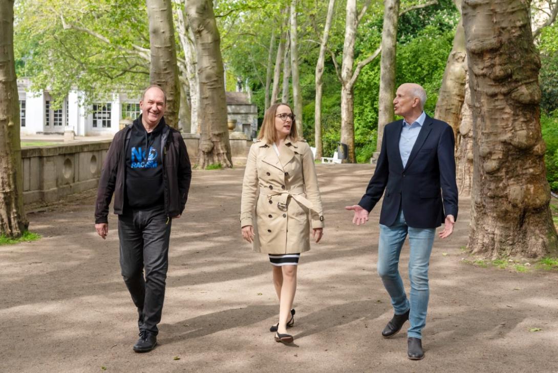 Gruppenbild von Gerd Thomas, Marcia Behrens und Ulrich Misgeld, während sie gestikulierend und sich angeregt unterhaltend durch einen Park spazieren.